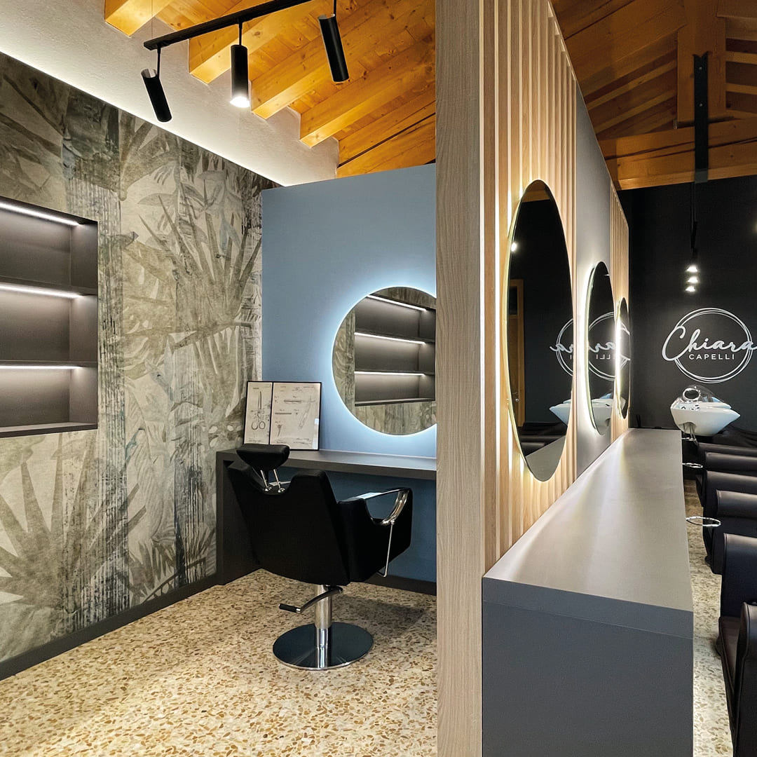 Immagine di una postazione dell'area stilistica con pareti azzurre alternate da pareti neutre con motivi floreali. Scaffali a muro illuminati e specchio rotondo 